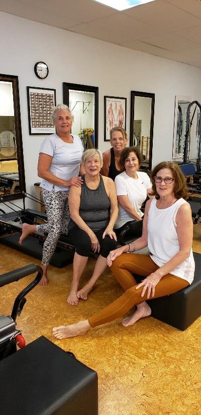Five smiling clients sitting together on reformers after class